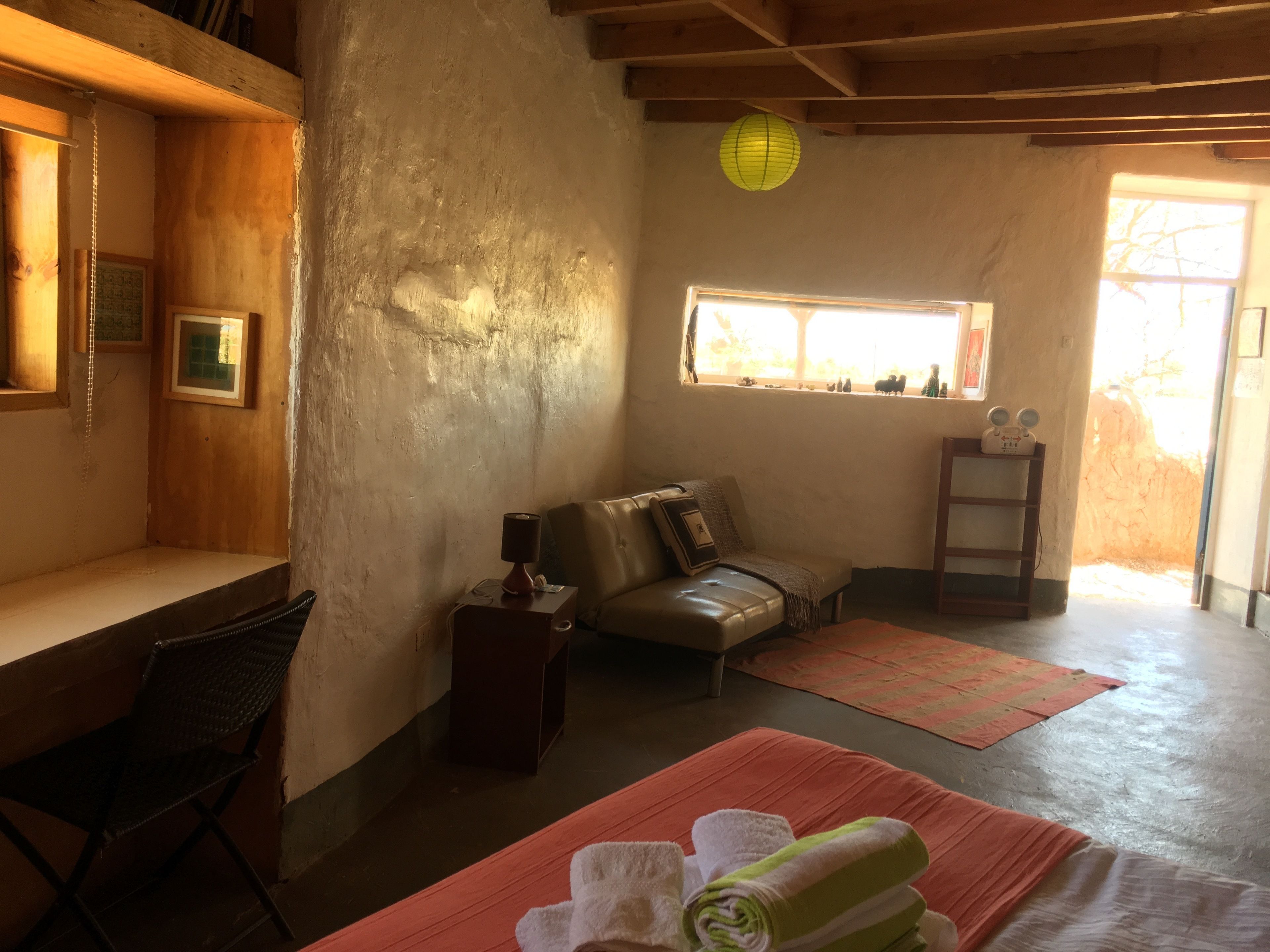 A cozy room with a wooden bench, a cushioned bench, a small table with a lamp, a window with a view, and a shelf with decorative items.
