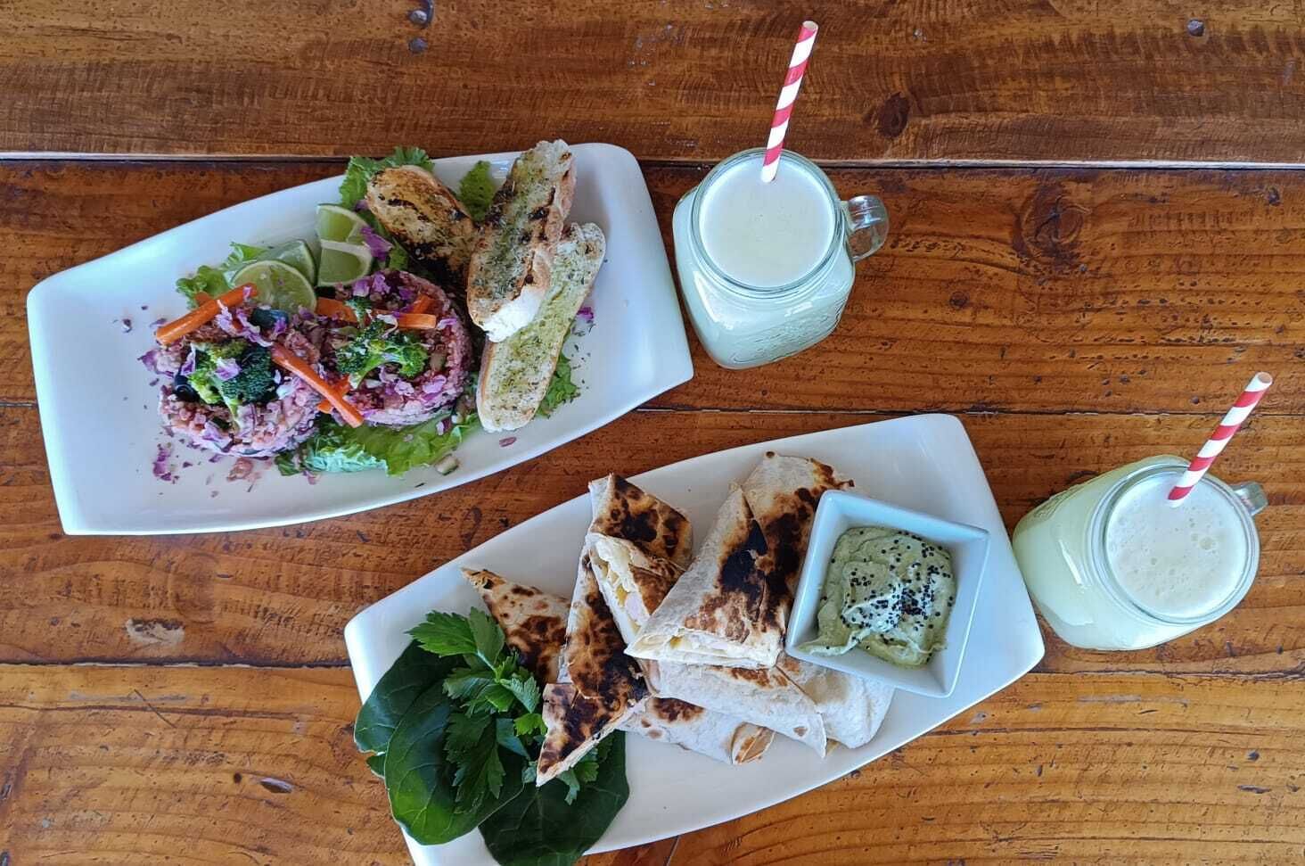 Uma mesa de madeira com dois pratos de comida, um com salada e outro com grelhados e uma porção de guacamole. Também há dois copos de uma bebida clara com canudos.
