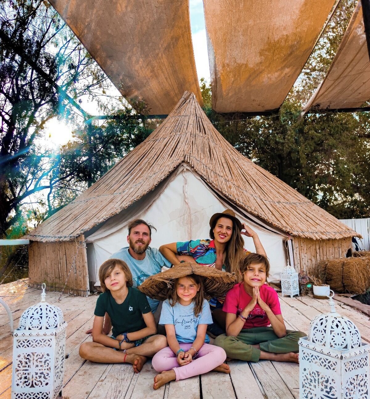 A family of five sitting in front of a thatched-roof hut with white lanterns on either side.