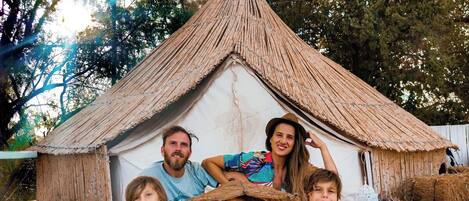 A family of five sitting in front of a thatched-roof hut with white lanterns on either side.