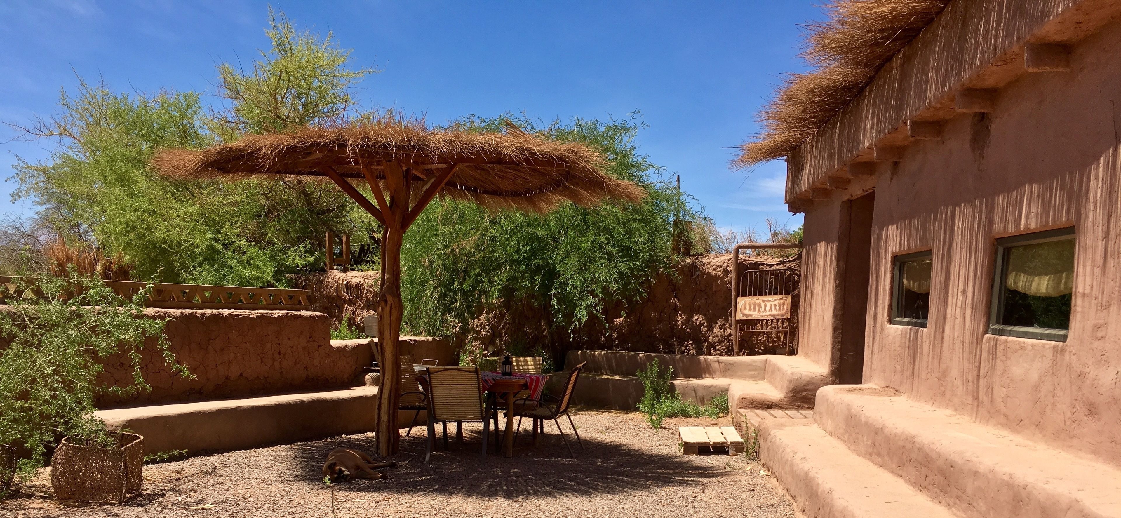 Un área de estar rústica al aire libre con techo de paja, rodeada de vegetación natural y una edificación tradicional.