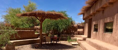 Un área de estar rústica al aire libre con techo de paja, rodeada de vegetación natural y una edificación tradicional.