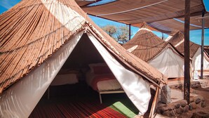Una tienda tradicional con techo de paja, un área de descanso con colchoneta a rayas rojas y un muro de piedra que rodea el campamento.