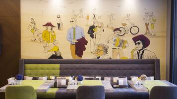 Un restaurant dont un mur est décoré d’une fresque représentant des personnes, avec un banc recouvert d’un tissu vert et des tables dressées avec des assiettes blanches et des fleurs bleues.