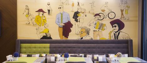 Un restaurante con un mural de gente en la pared, un banco tapizado en verde y mesas dispuestas con platos blancos y flores azules.
