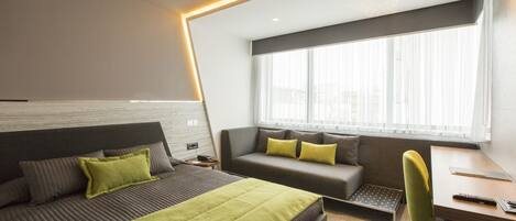 A modern hotel room with a bed, a sofa, a desk, and a window with curtains.