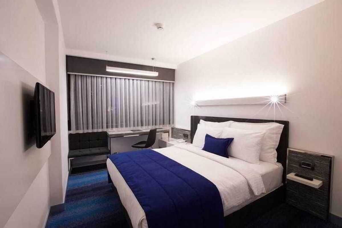 Habitación de hotel con cama, escritorio, silla, televisión y ventana con cortinas.