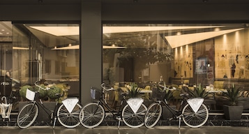Una hilera de bicicletas estacionadas frente a un edificio con puertas de vidrio, un área de descanso con mesas y sillas, y una planta grande al fondo.