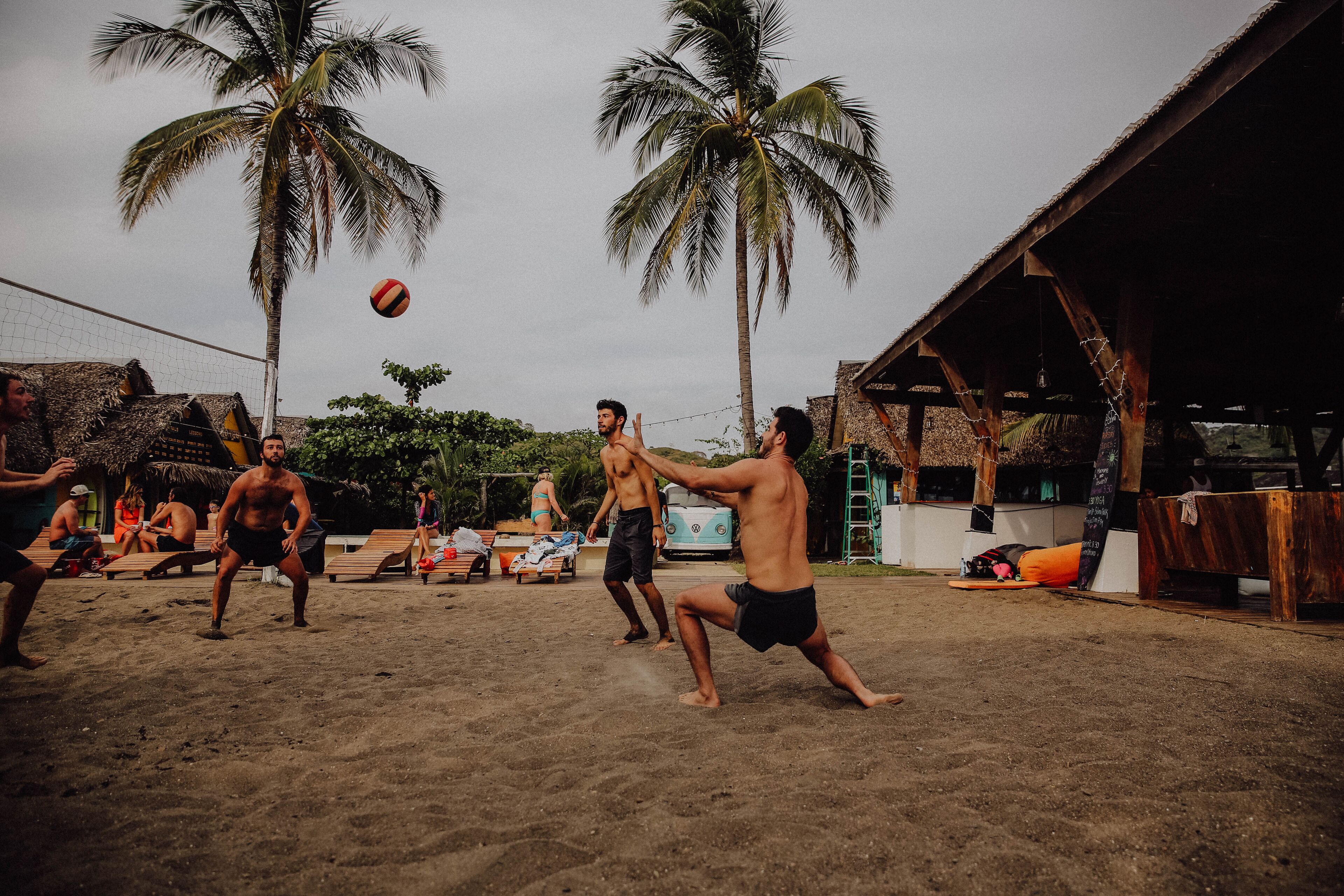 Partita di beach volley su una sabbiosa spiaggia, con palme e una struttura in legno sullo sfondo.