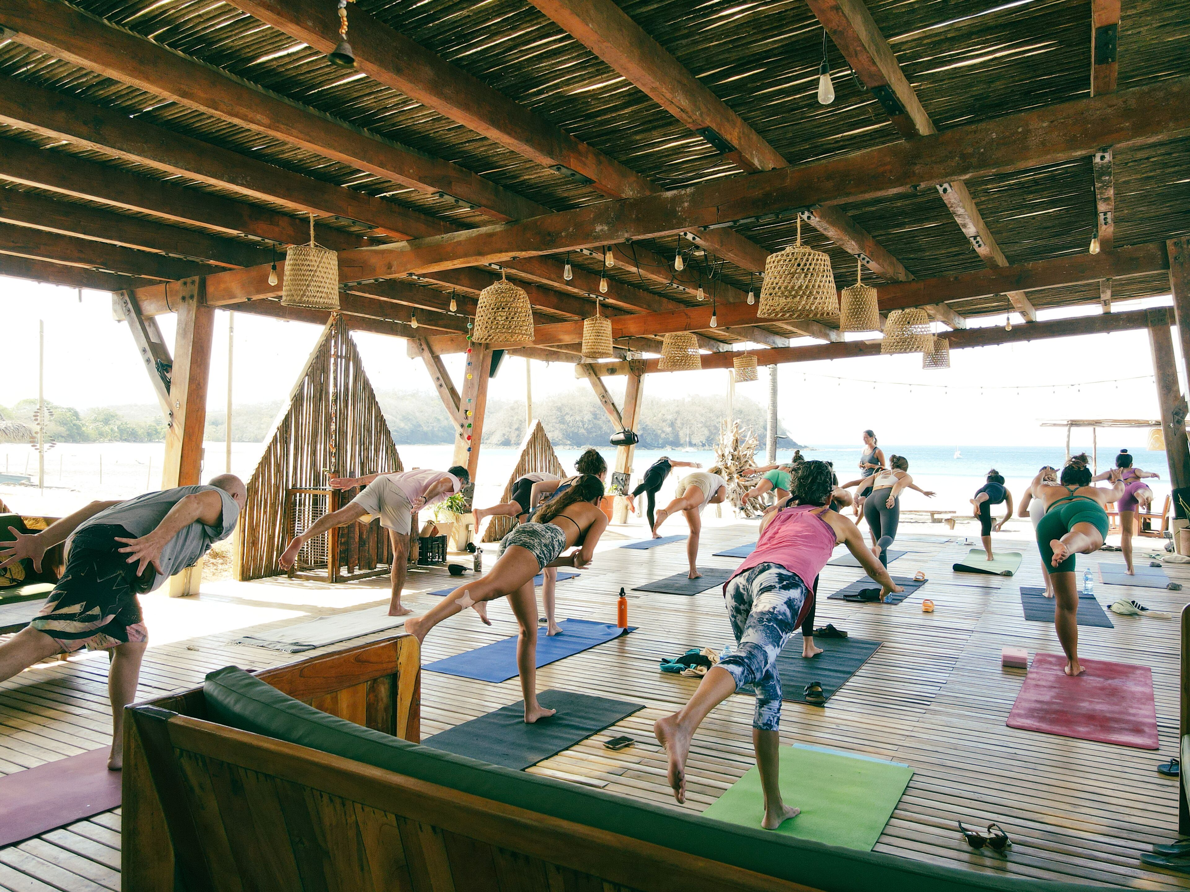 Un grupo de personas practicando yoga en colchonetas en una terraza de madera al aire libre con vista al mar.