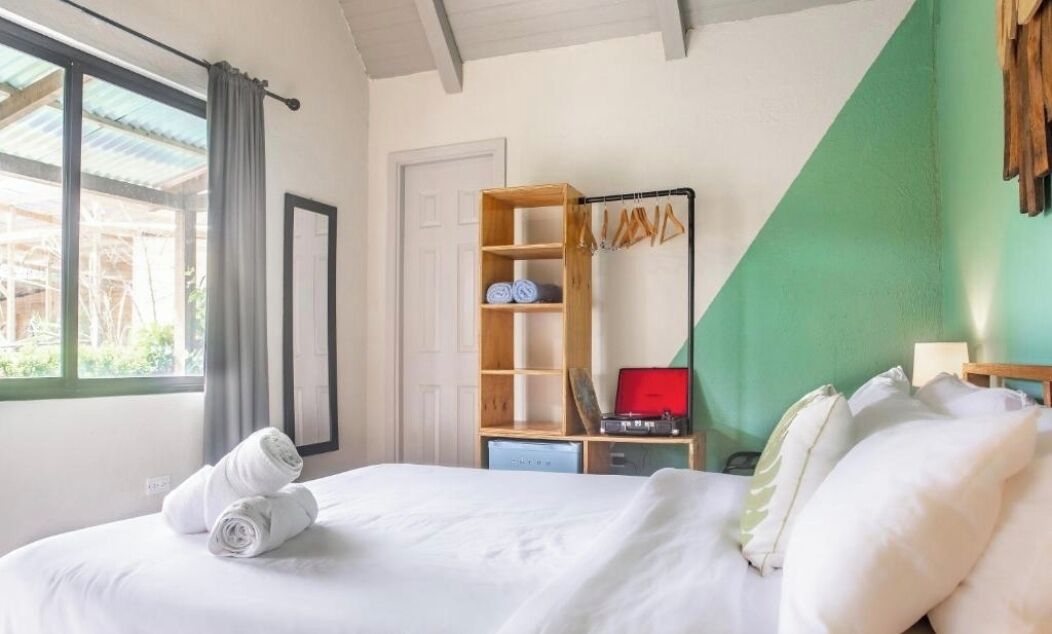 A hotel room with a bed, a wooden wardrobe, a desk with a red suitcase, and a window with curtains.
