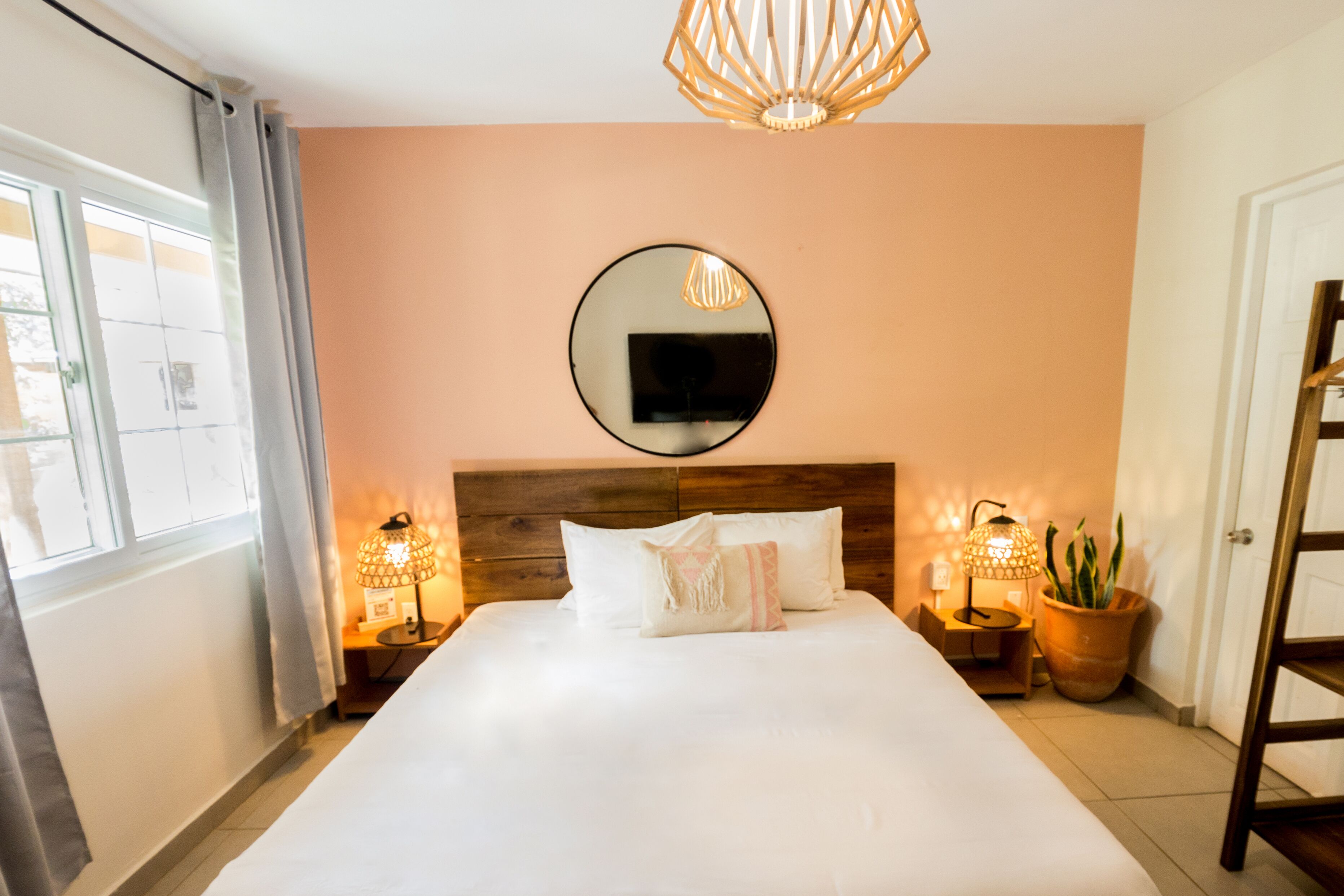 A neatly made bed with white linens, a round mirror on the wall, two bedside lamps, and a potted plant.