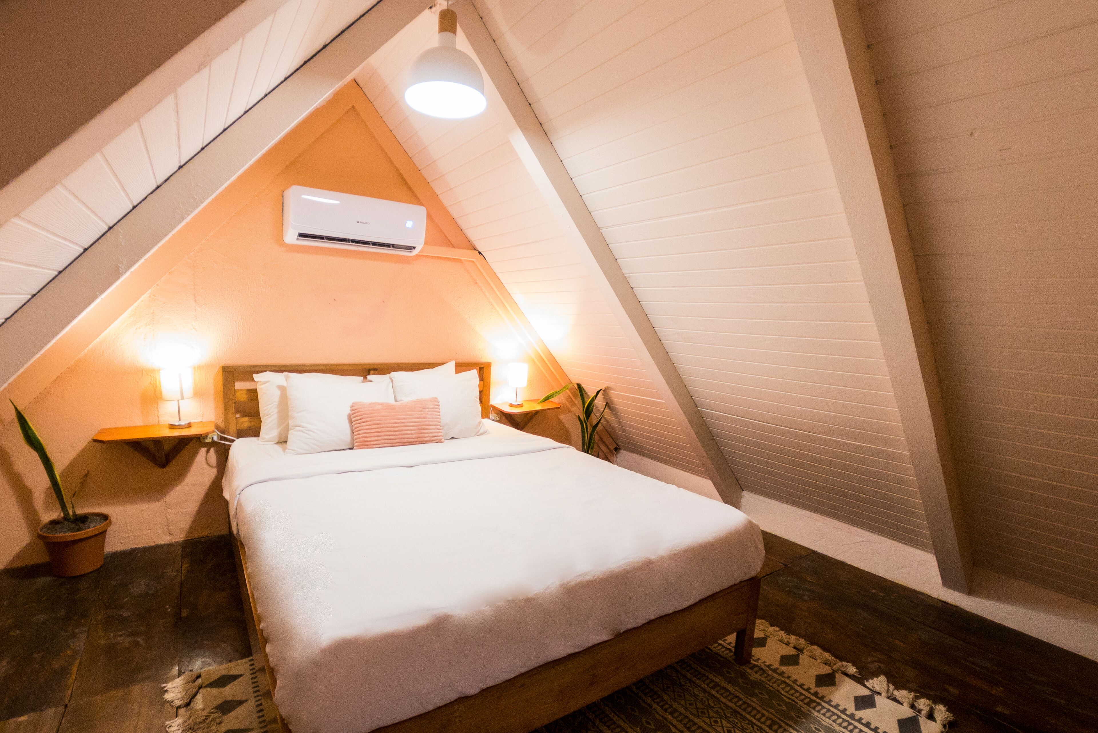 A bedroom with a sloped ceiling, a bed with white bedding, two bedside lamps, a potted plant, and an air conditioning unit.