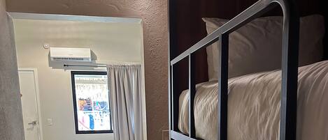 Hotellihuone, jossa on kerrossänky, yhden hengen sänky kuviollisilla vuodevaatteilla, ikkuna verhoilla ja ilmastointilaite.