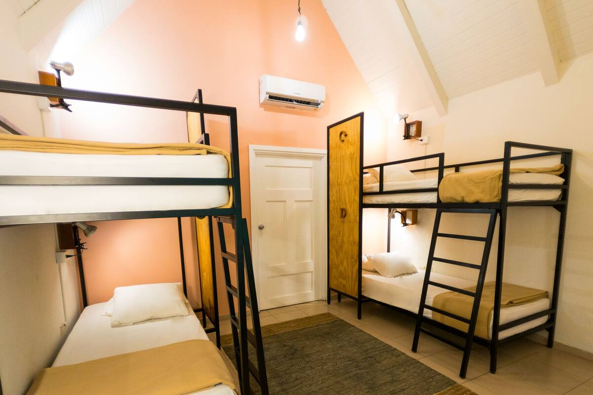 A dormitory room with bunk beds, a door, an air conditioning unit, and a small desk with a lamp.