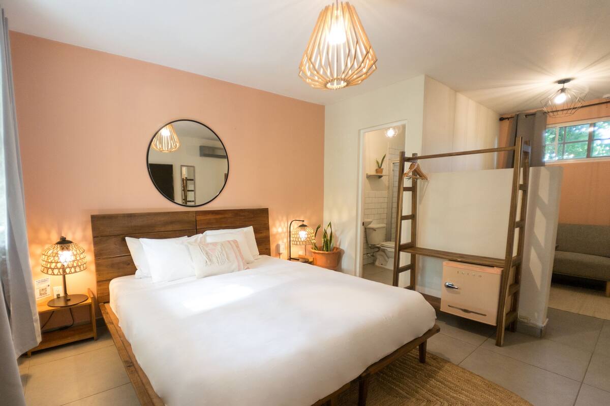 A bedroom with a bed, bedside lamps, a mirror, a hanging light fixture, a wardrobe, and a bathroom visible through an open door.