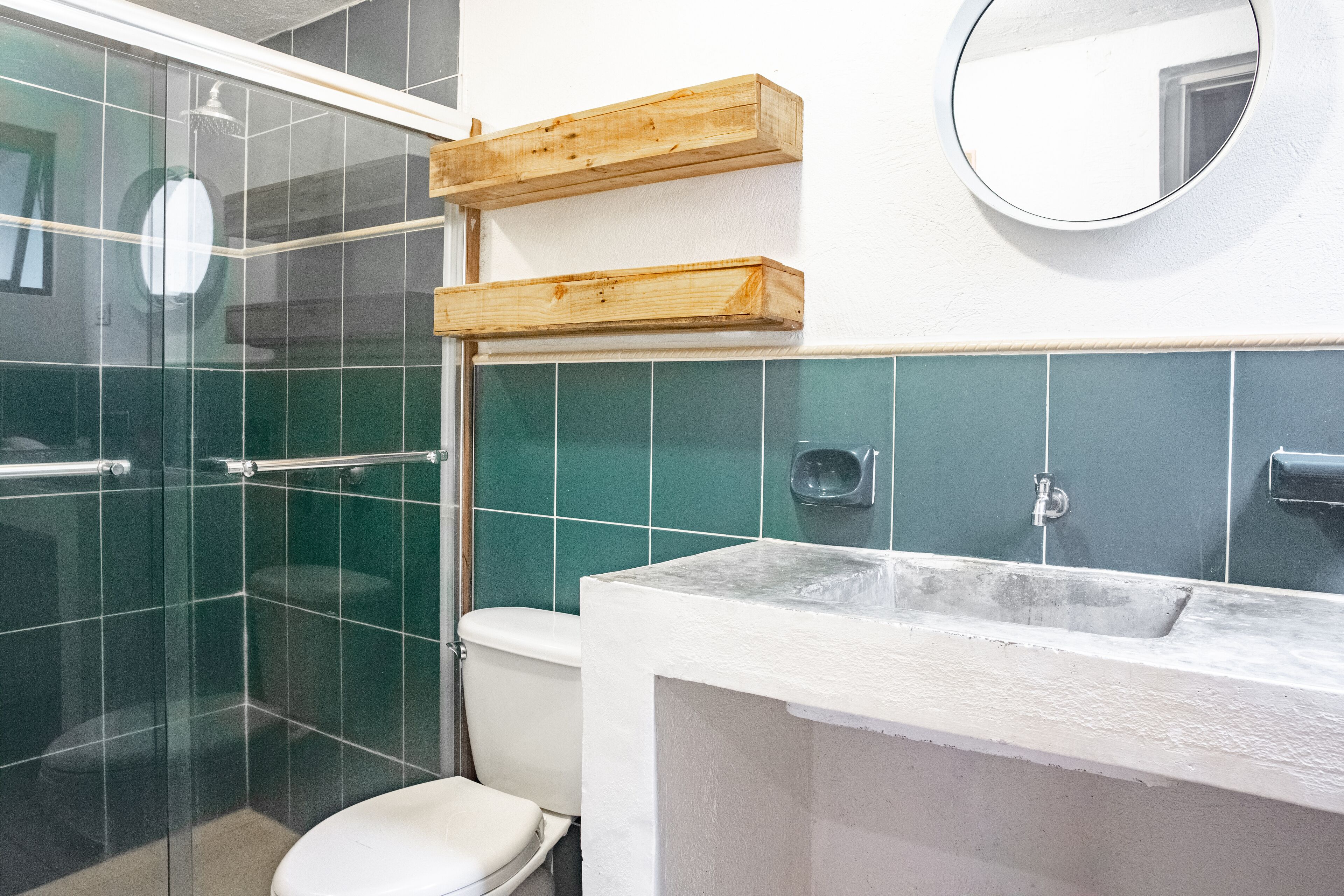 Un baño con cabina de ducha de vidrio, inodoro, lavamanos con encimera blanca y estantes de madera sobre el lavamanos.