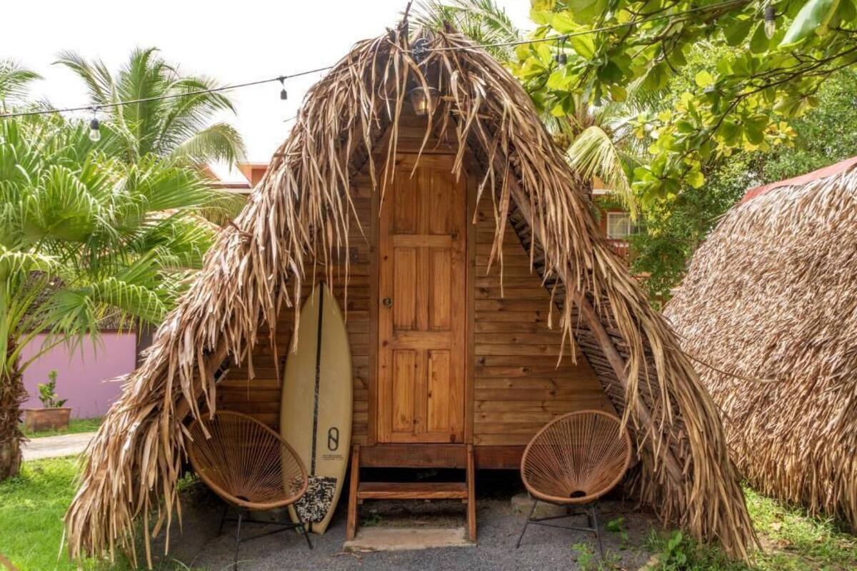 A thatched-roof hut with a wooden door, two wicker chairs, and a surfboard leaning against the door.