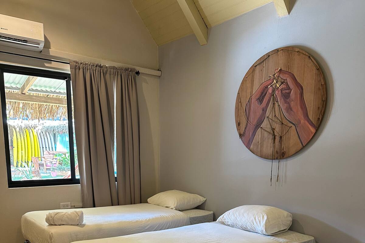 A hotel room with three beds, a window with curtains, an air conditioner, and a wall-mounted artwork.