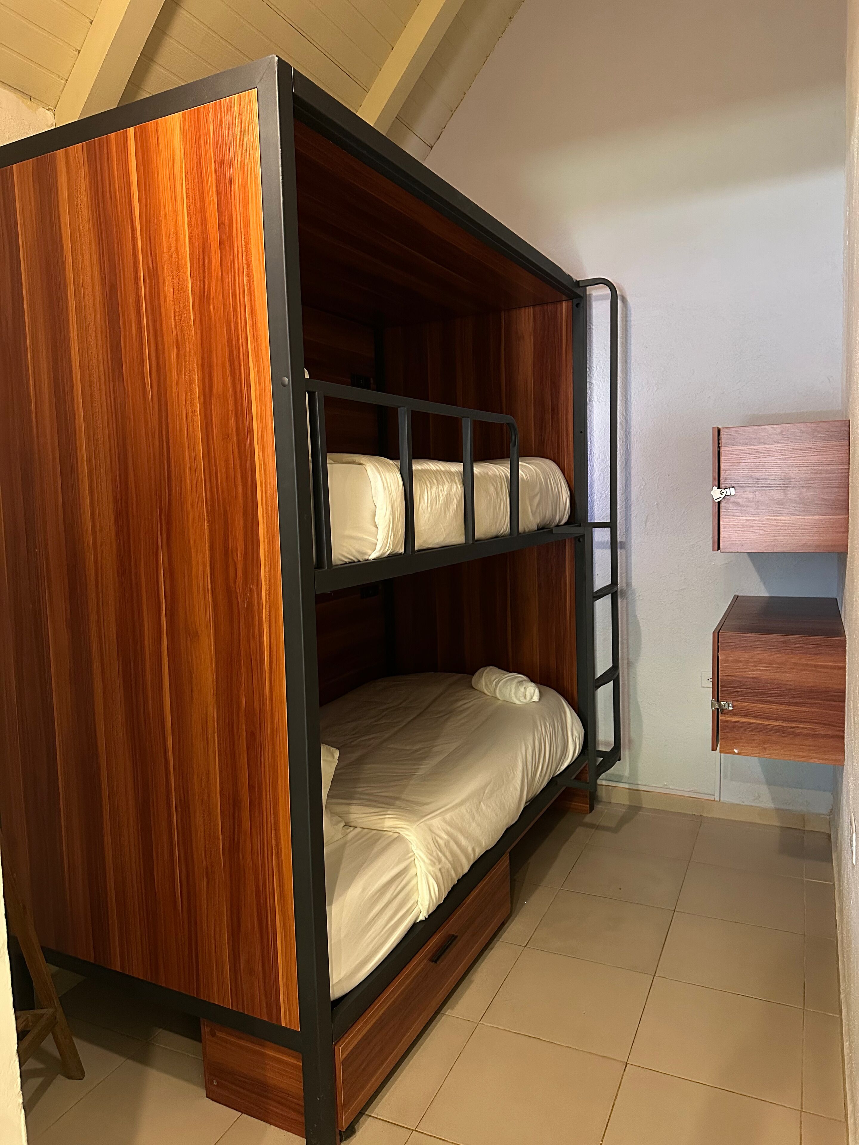 A bunk bed with a wooden finish and a metal frame, a small wooden cabinet, and a tiled floor.