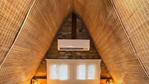 A cozy, triangular-roofed bedroom with a wooden bed, a white mattress, and a pillow. There is a window with a view of the outdoors, and a wall-mounted air conditioning unit above the bed.