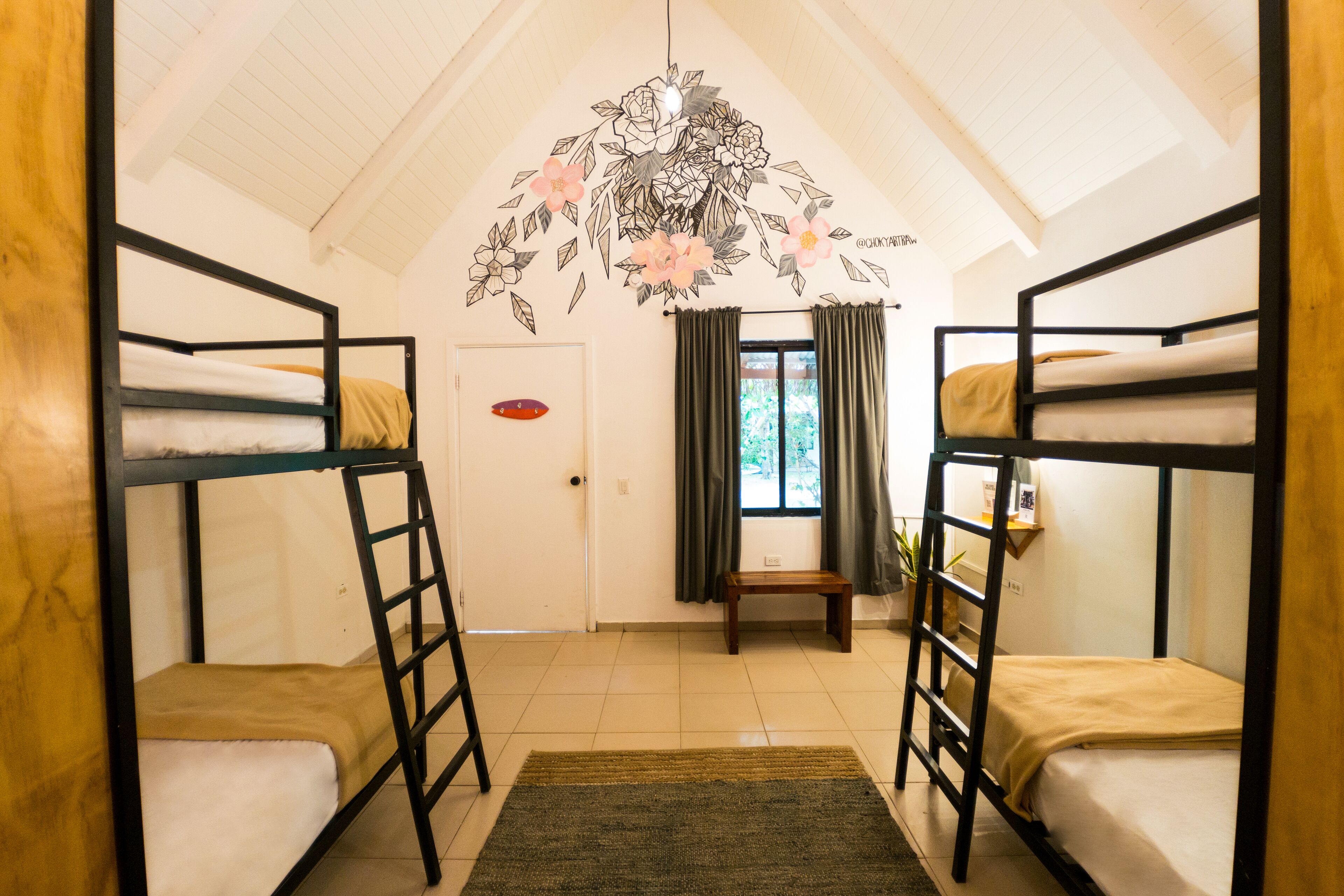 A dormitory room with bunk beds, a wooden door, a small bench, and a floral wall decoration.