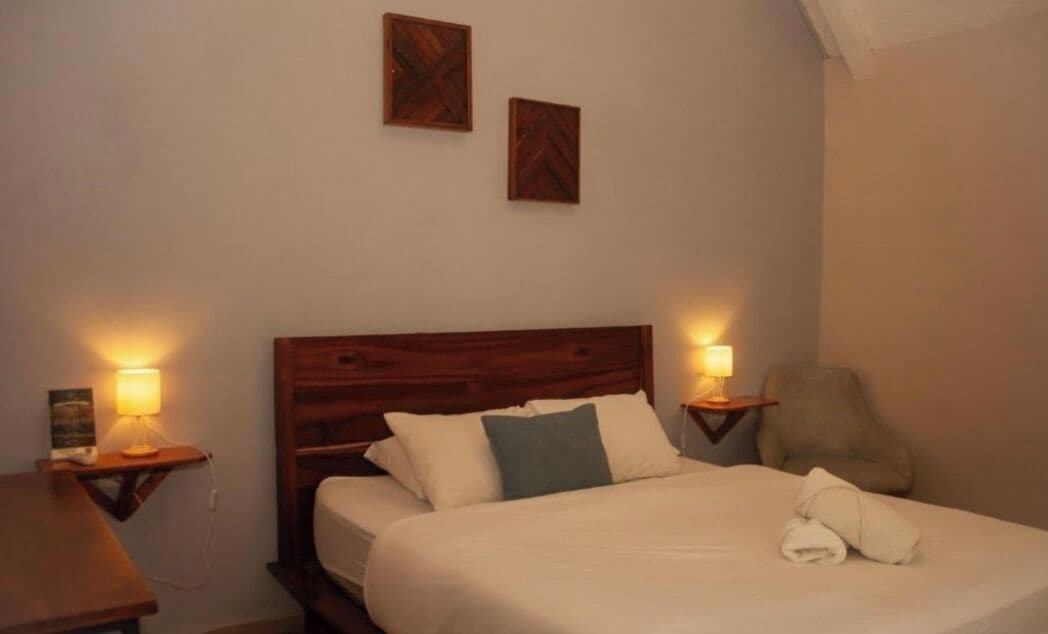A neatly made bed with pillows, a wooden headboard, two bedside lamps, and a chair.