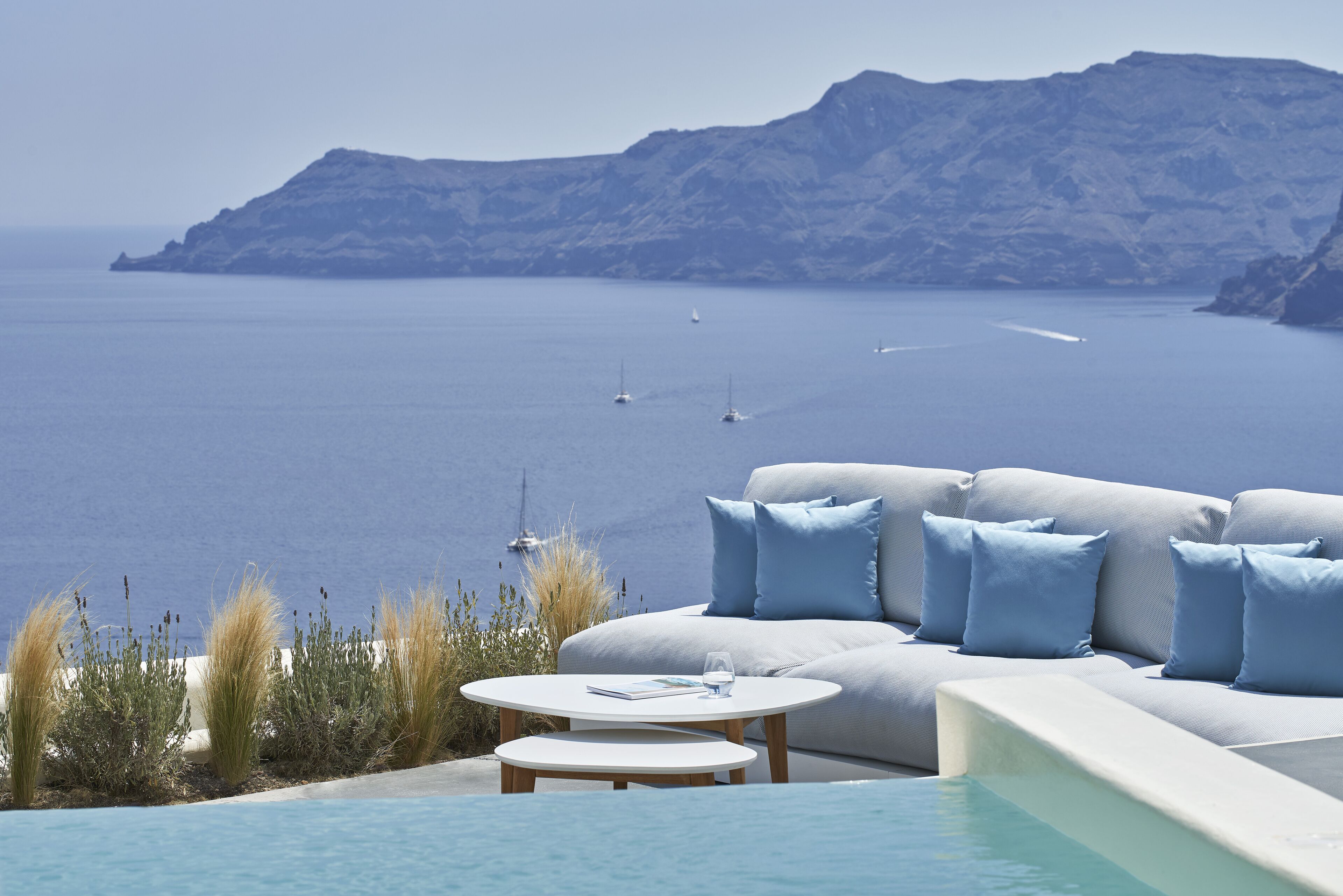 Ein moderner Außenbereich mit Sitzgelegenheit und Pool, mit Blick auf ein ruhiges Meer mit Booten und fernen Bergen.