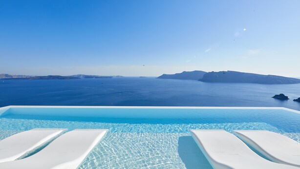 Una piscina infinita con agua azul cristalina y sillas de descanso blancas, con vista al mar tranquilo y a islas distantes.