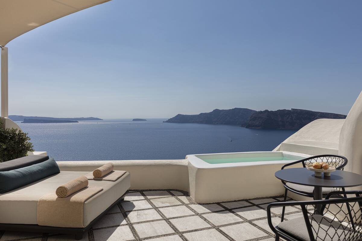 A balcony with a view of the sea, a sofa, a table with a bowl of fruit, and a small pool.