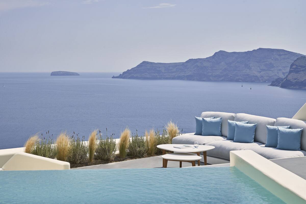 A poolside area with a sofa and a round table, overlooking a body of water and a mountainous landscape.