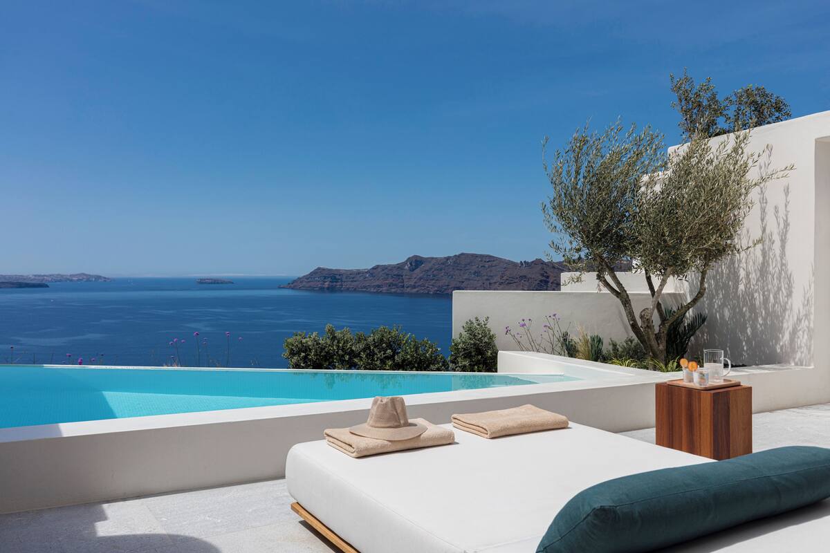 A modern outdoor lounge area with a pool, a white sofa, a wooden side table, and a view of the sea and distant land.