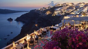 Un espacio de comedor frente al mar con mesas y sillas, con vistas a una bahía con barcos y un pueblo al fondo.