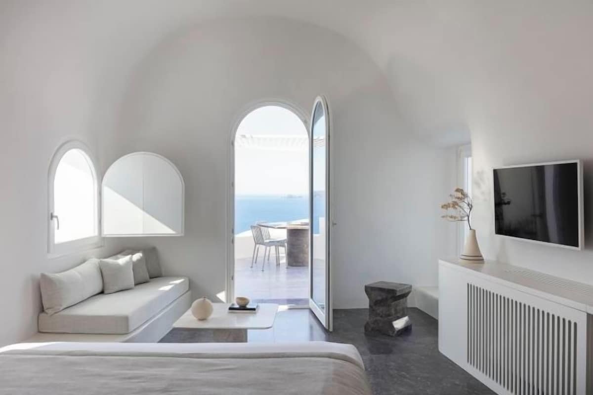 Un dormitorio moderno con cama, zona de estar con cojines, televisor y vista al mar.