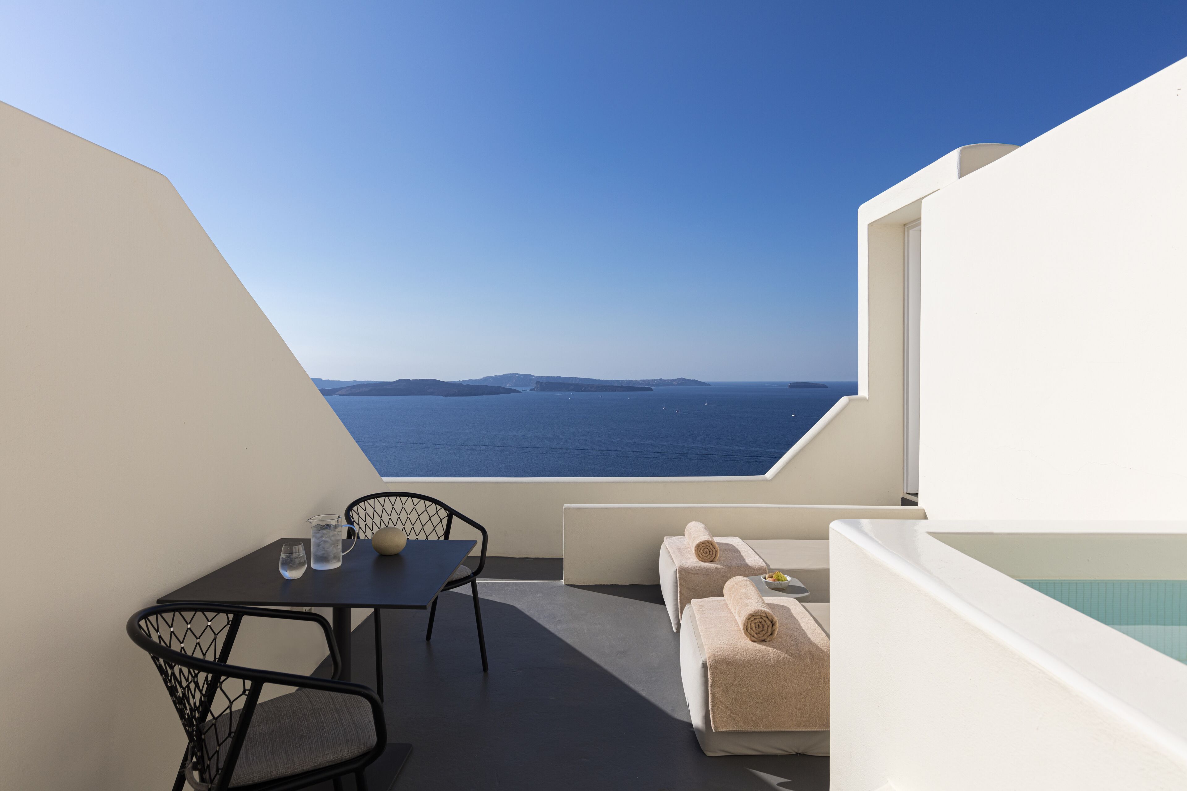 Un balcon avec une table et des chaises, un canapé avec des serviettes, et une vue sur la mer et les îles.