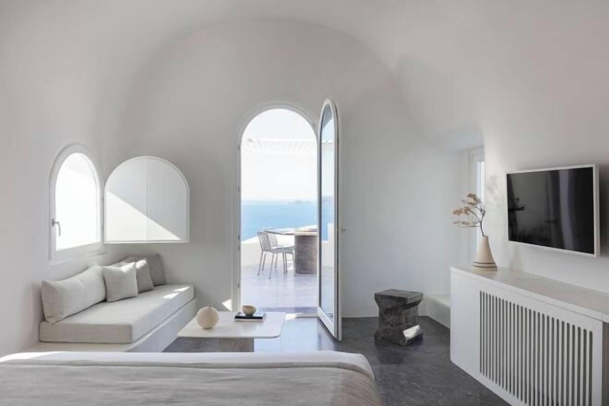 A modern bedroom with a bed, a seating area with cushions, a TV, and a view of the sea.
