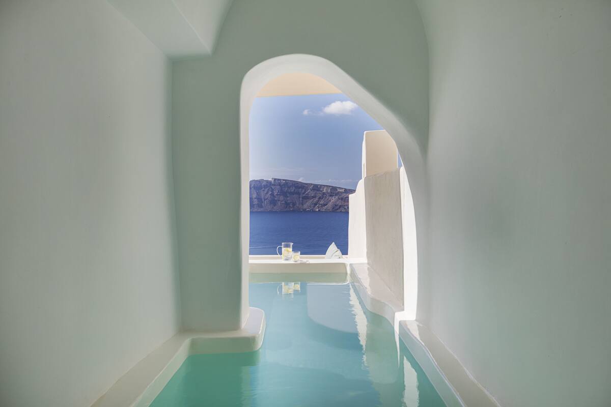A narrow, arched passage leading to a pool with a view of a cliff and the sea.