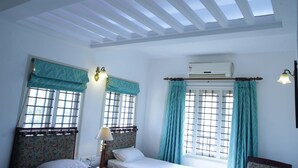 Una habitación con tragaluces, dos ventanas con cortinas, una cama con cabecera floral y un equipo de aire acondicionado.