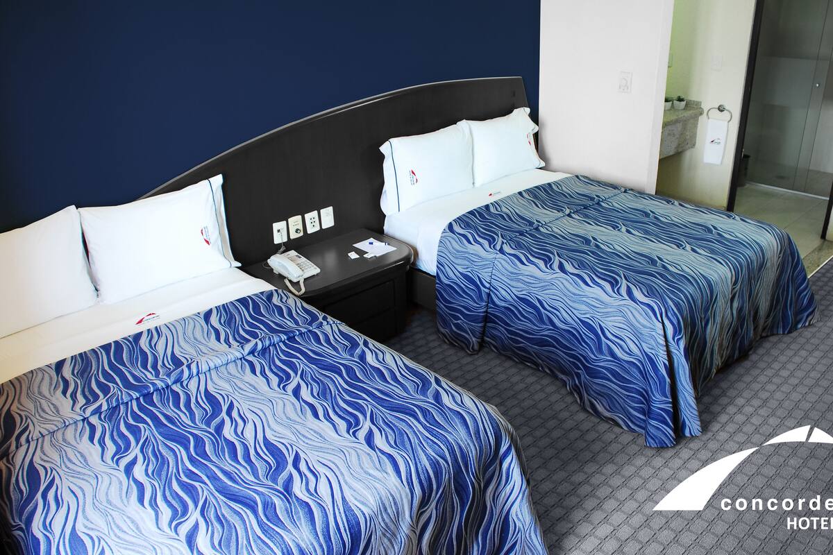 A hotel room with two beds, each with a blue patterned bedspread, a bedside table with a phone, and a nightstand.