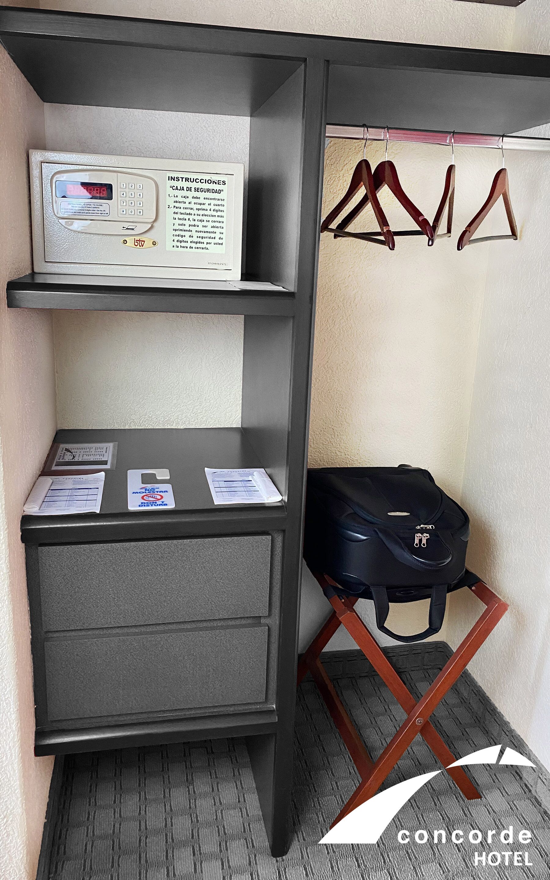 Una habitación de hotel con un armario gris, una caja fuerte, una maleta negra sobre un taburete de madera y un perchero con ropa.