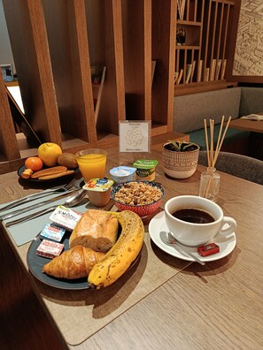 A breakfast table with croissants, a banana, a bowl of granola, a glass of orange juice, a cup of coffee, and a small sign reading "Bienvenue". -  Lyo Hôtel (Lyon)