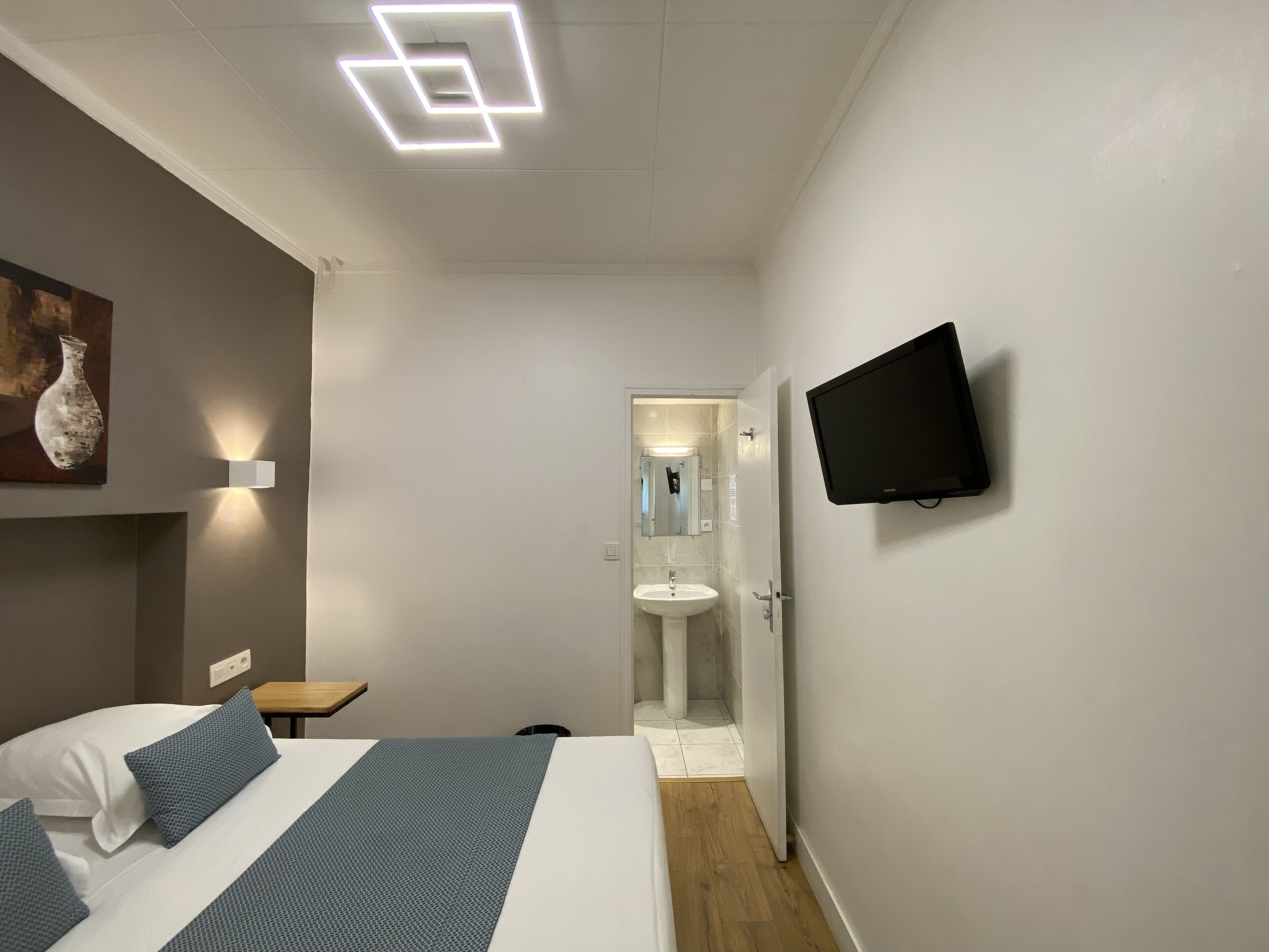 A modern hotel room with a bed, bedside table, wall-mounted TV, and a bathroom visible through an open door.