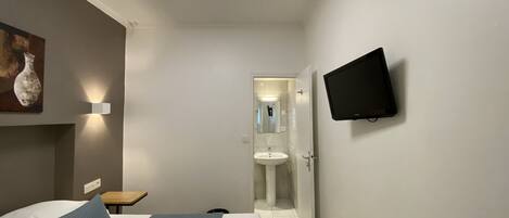 A modern hotel room with a bed, bedside table, wall-mounted TV, and a bathroom visible through an open door.