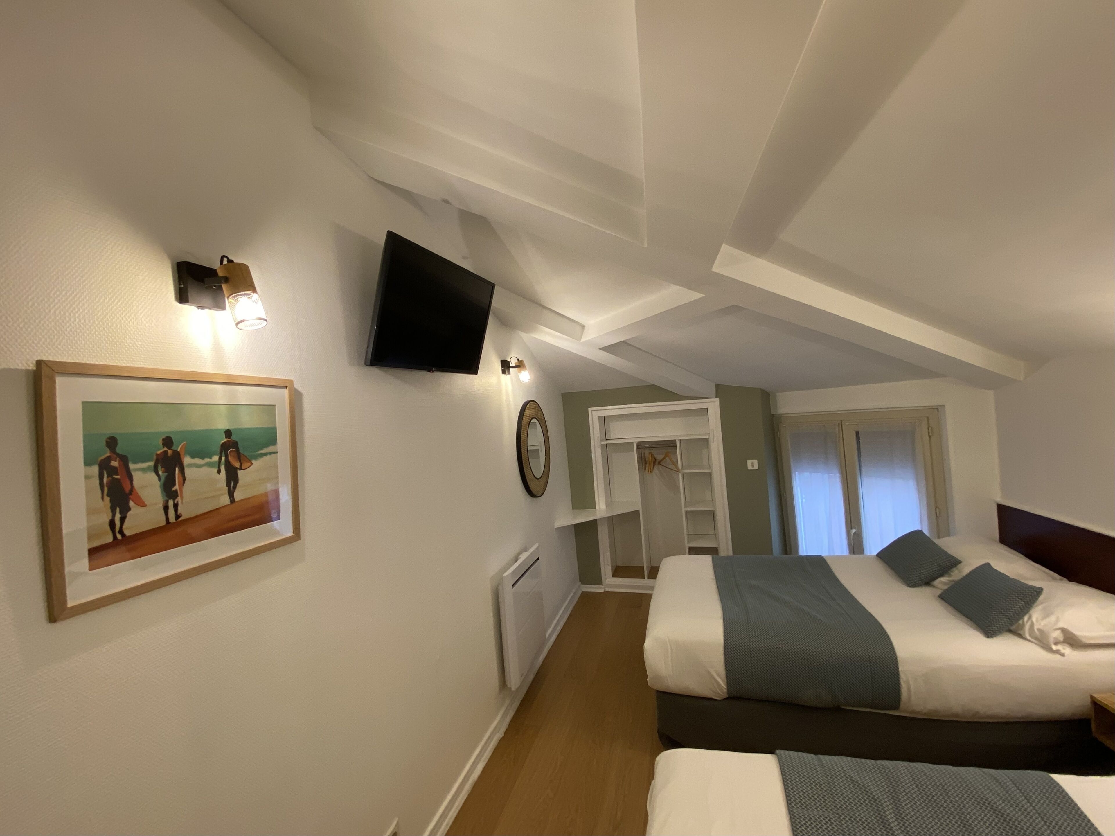 A hotel room with two beds, a television mounted on the wall, a framed picture of surfers, and a walk-in closet.
