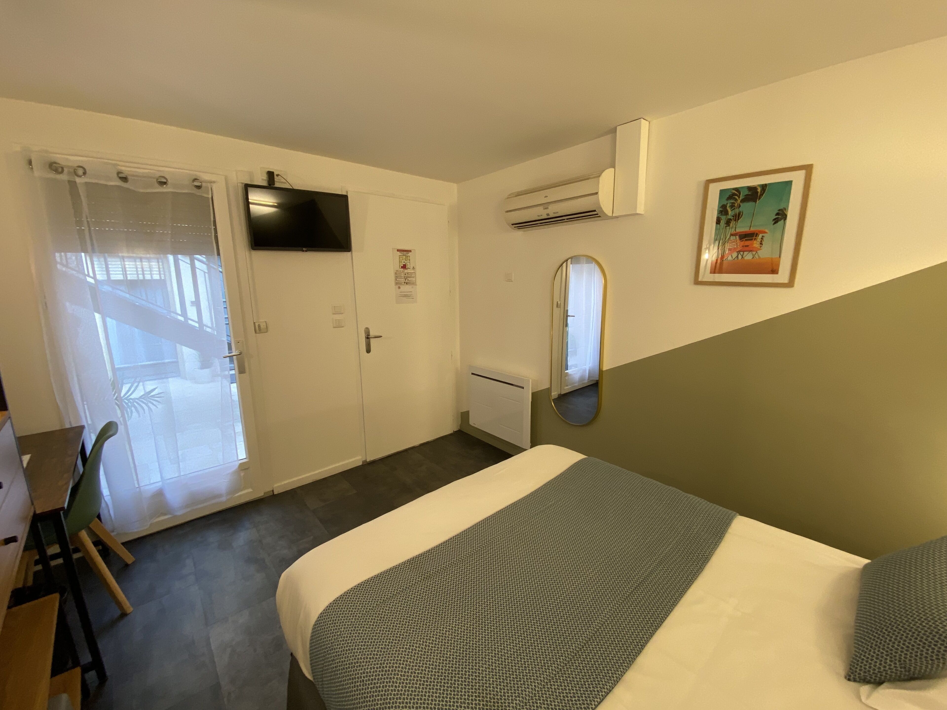 A hotel room with a bed, a desk, a chair, an air conditioning unit, a mirror, and a door with a frosted glass window.
