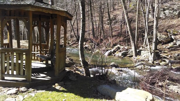 Un belvédère en bois avec un banc, entouré d’une zone boisée traversée par un ruisseau.