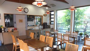 A restaurant with wooden tables and chairs, a kitchen area, and large windows with a view of greenery.