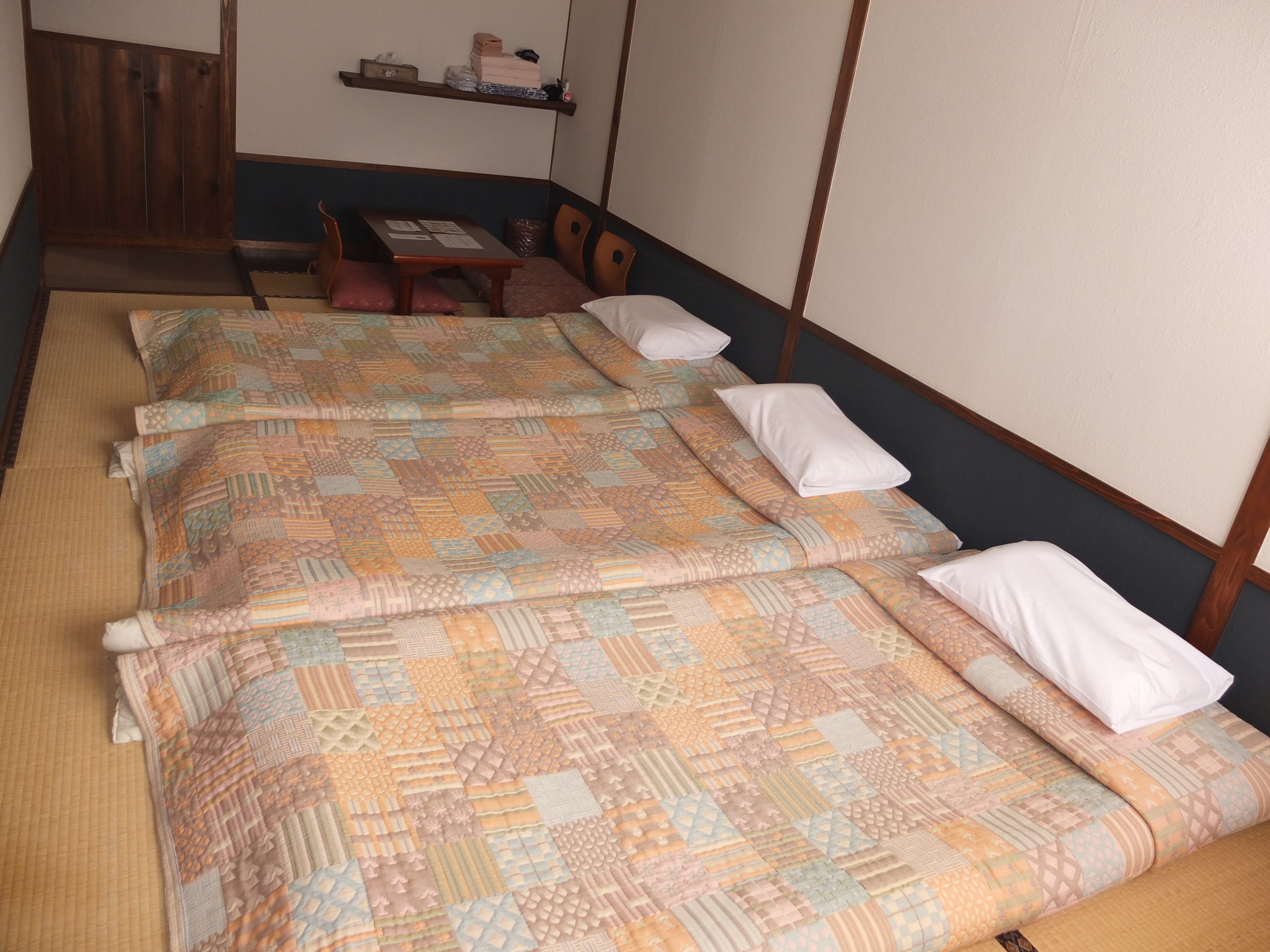 A traditional Japanese room with tatami flooring, a low wooden bed with a patterned quilt, a small table with a cushion, and a shelf with personal items.