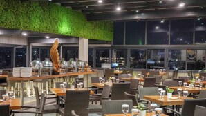 A restaurant with tables set for dining, a green wall, and city views through large windows.