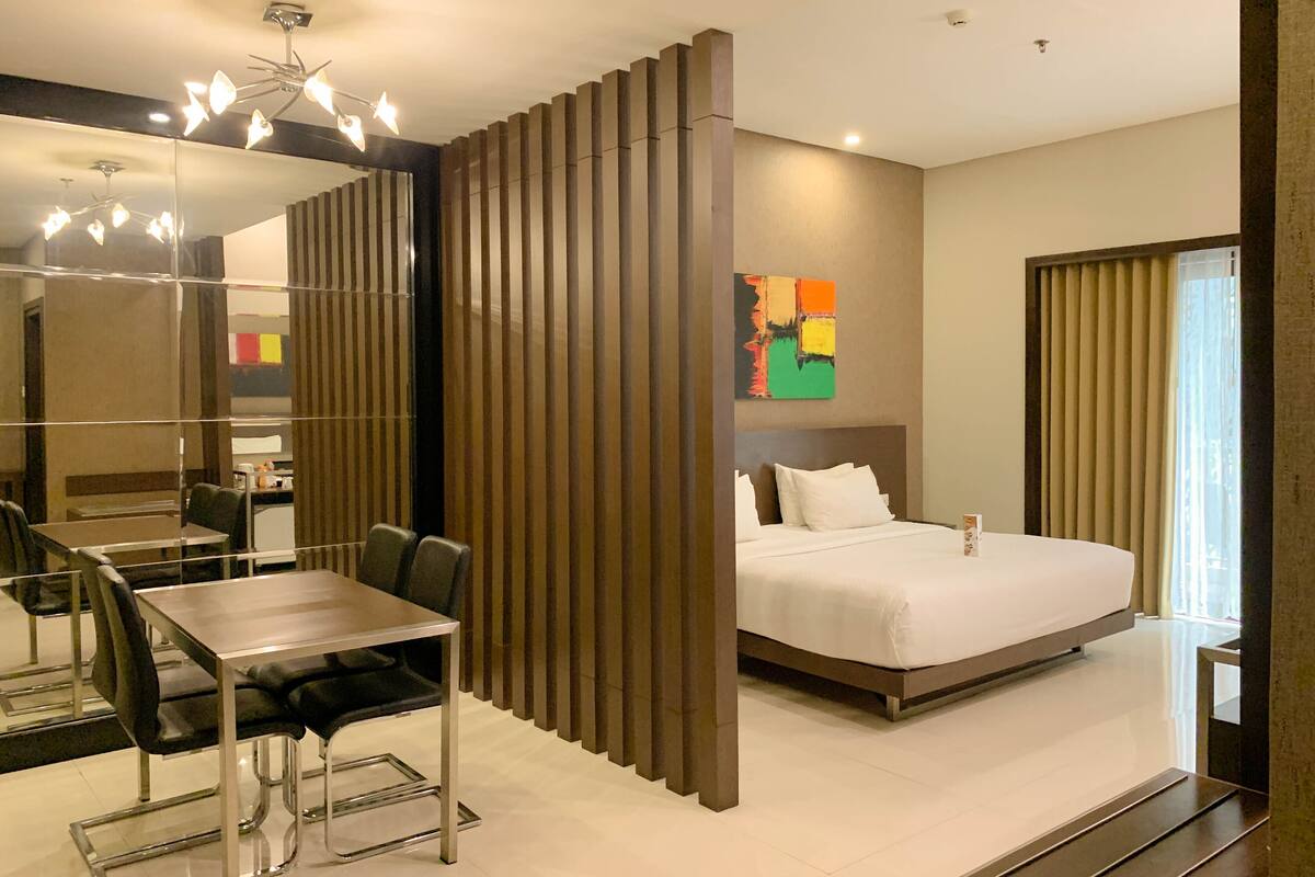 A modern hotel room with a bed, a desk with chairs, a large mirror, and a colorful abstract painting on the wall.